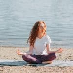 A woman practising meditation and yoga on the beach by the calm water, promoting mindfulness and relaxation.